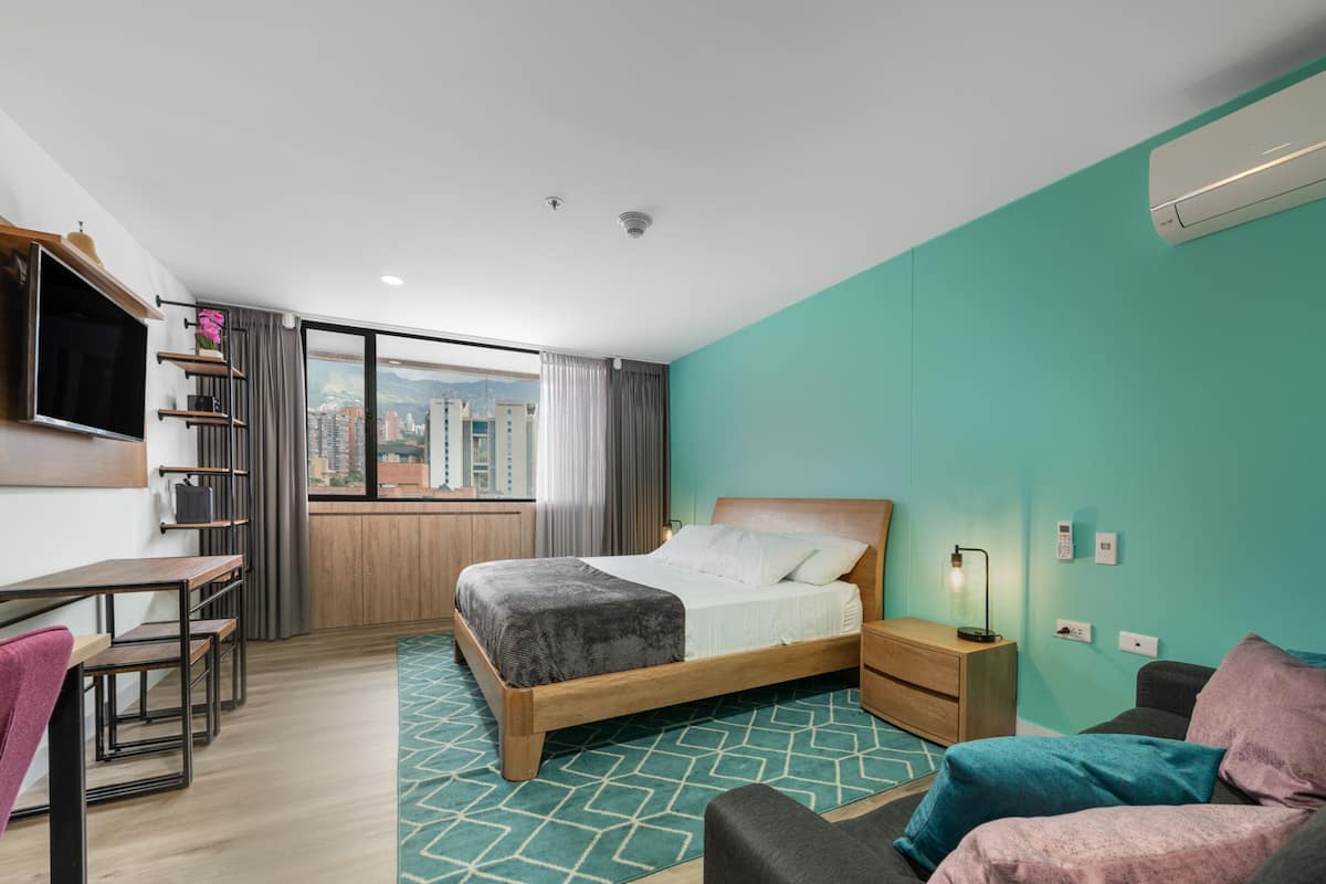 Una habitación de hotel moderna con cama, escritorio, silla, sofá y vista de la ciudad a través de amplias ventanas.