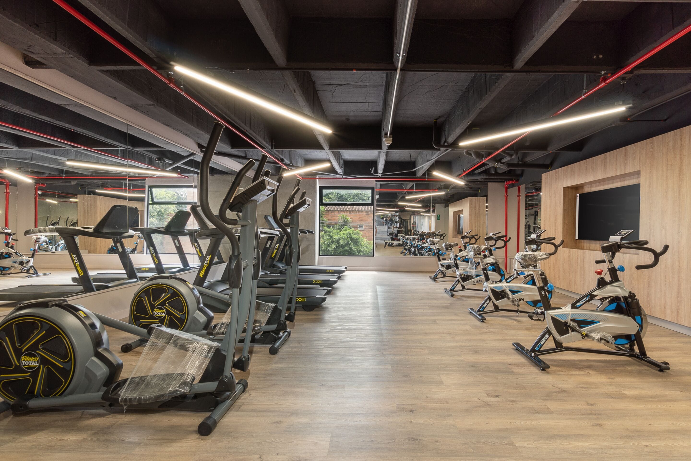 A gym with multiple treadmills and stationary bikes, wooden flooring, and large windows offering a view of greenery.