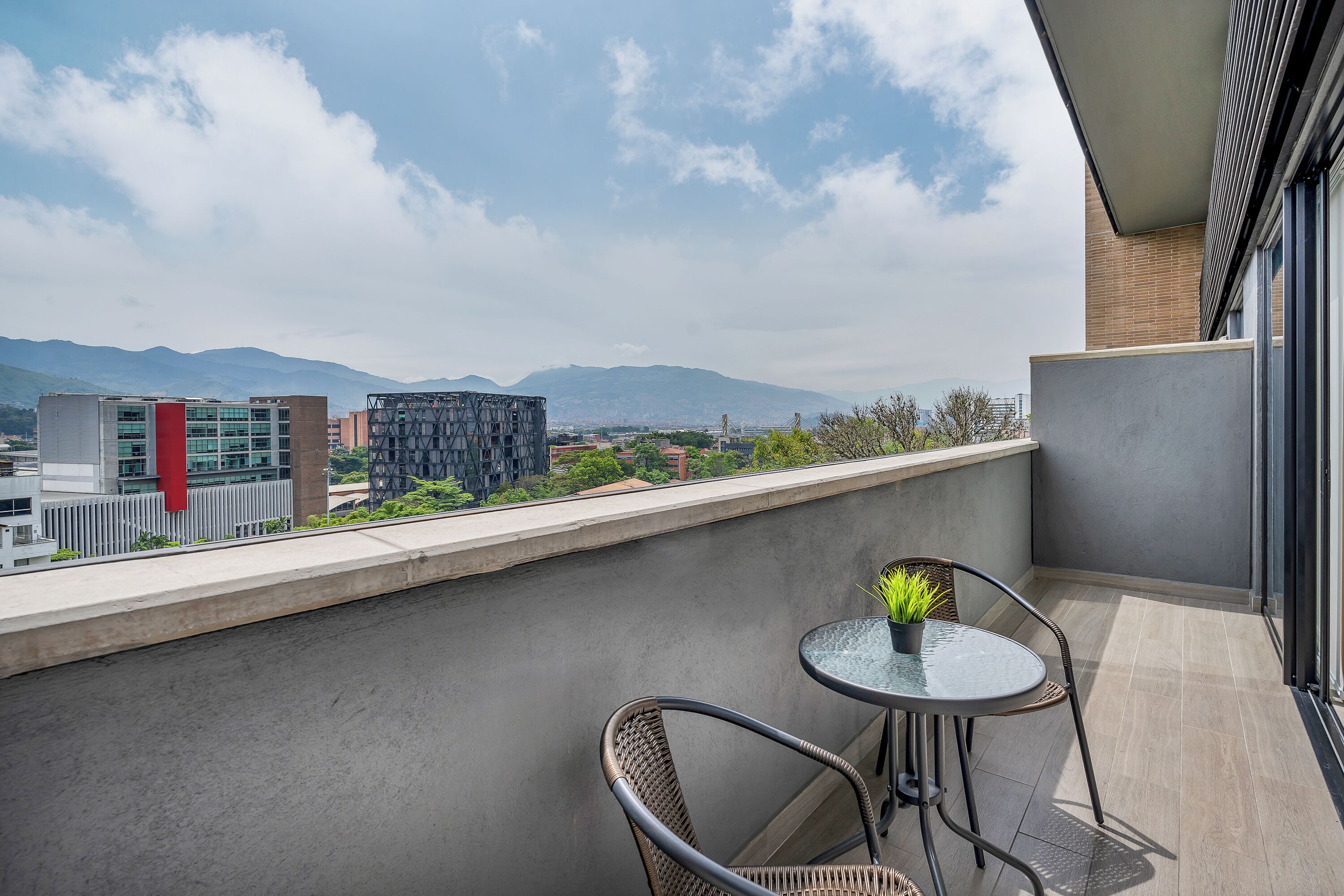 A balcony with a view of mountains, a table with a plant, and wicker chairs.