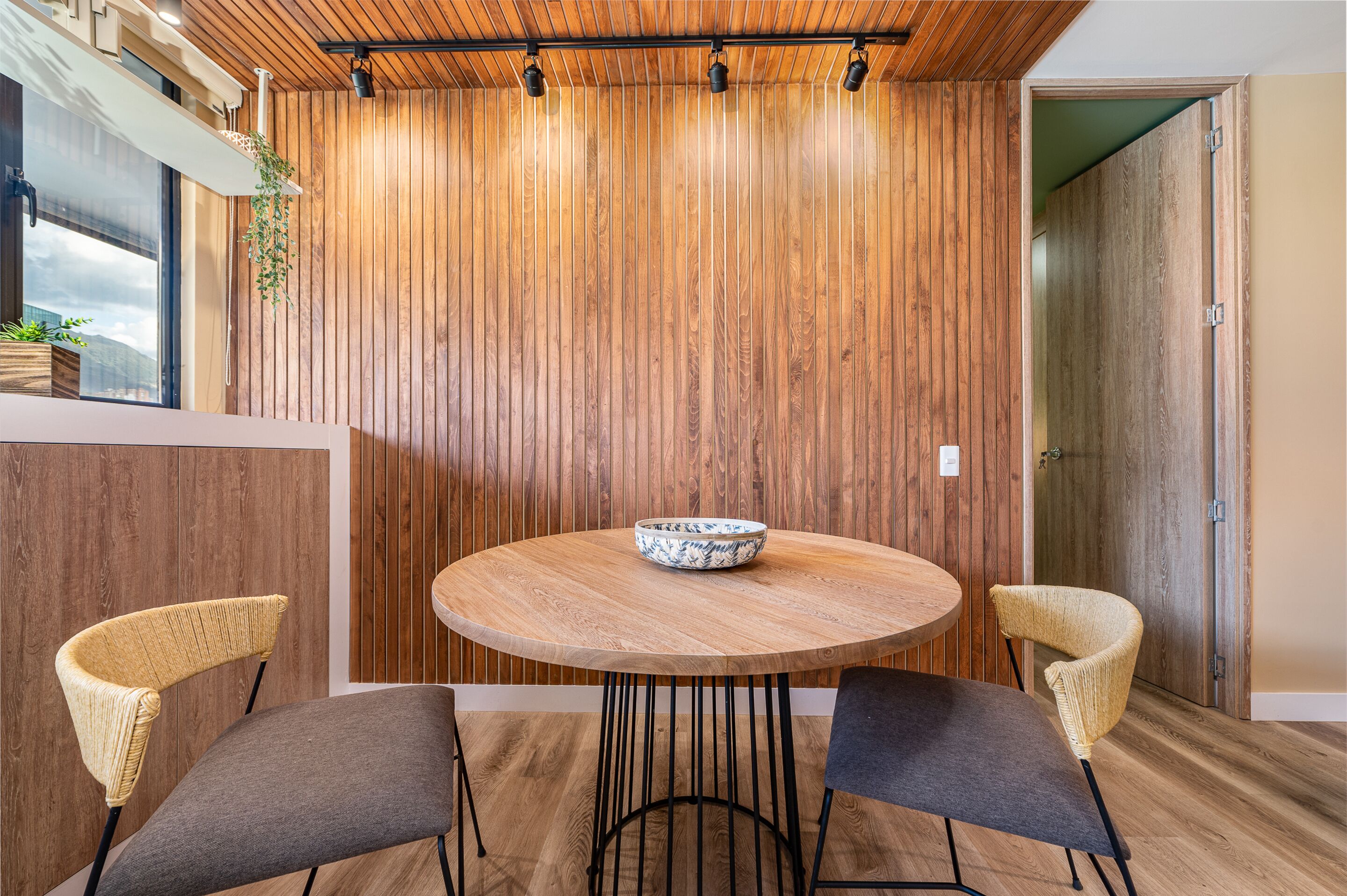 Een moderne eetruimte met een ronde houten tafel, twee rieten stoelen en een houten wand met verticale latten.
