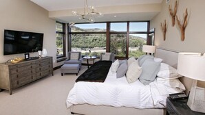 Ein Schlafzimmer mit einem großen Bett, einem Fernseher, einer Kommode, einem Sessel und Blick ins Freie.