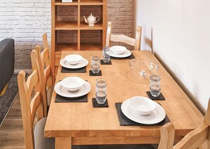 A wooden dining table set with plates, bowls, and glasses, surrounded by chairs, with a brick wall and a shelf in the background.