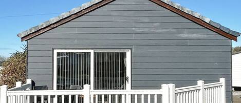 A small, single-story building with a gray exterior, a white porch with a railing, and a large sliding glass door.