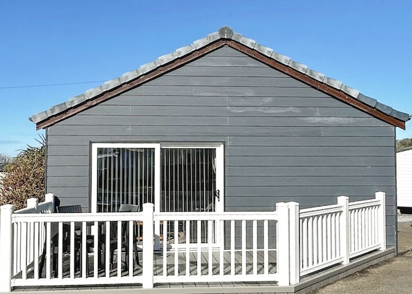 A small, single-story building with a gray exterior, a white porch with a railing, and a large sliding glass door.