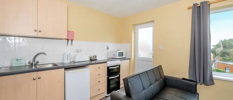 A compact kitchen with a sink, toaster, microwave, and a small refrigerator. There's a black leather sofa and a window with curtains.