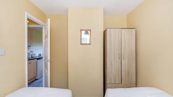 A small room with two beds, a wardrobe, a door leading to a kitchenette, and a small window.