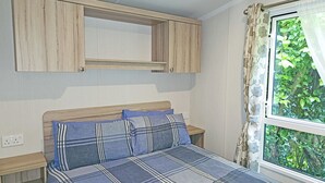 A bedroom with a bed, a wooden headboard, a window with curtains, and a built-in wooden shelf above the bed.