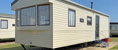 Une maison mobile de plain-pied, avec une entrée à l’avant, deux fenêtres avec des rideaux, un petit espace porche et une cheminée visible.