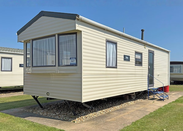 A single-story mobile home with a front entrance, two windows with curtains, a small porch area, and a visible chimney.