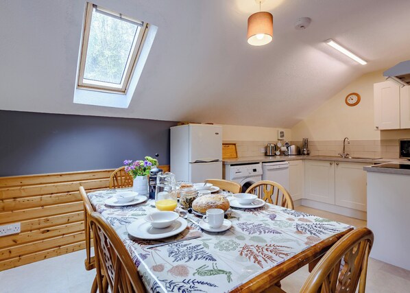 A dining area with a table set for four, a window, a ceiling light, and a kitchen in the background. - 2 bedroom accommodation in Webbery, Bideford (Webbery, Bideford)