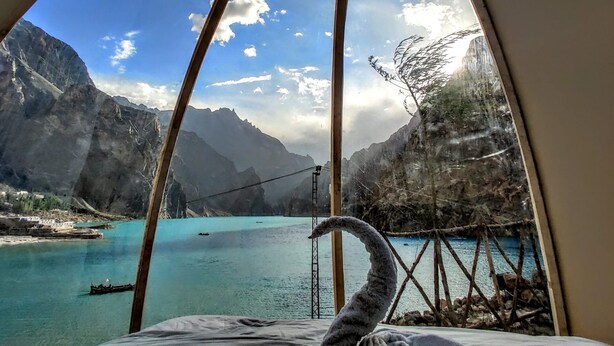 Uma tenda com uma janela circular que dá para um lago azul-turquesa, montanhas e uma ponte suspensa.
