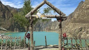 Um arco de madeira rústico com uma placa, rodeado por um espaço de cascalho e com vista para um lago azul-turquesa com montanhas ao fundo.