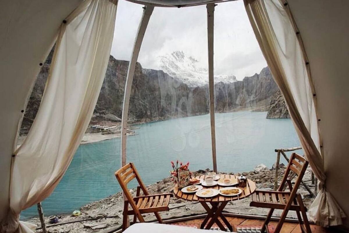 A cozy room with a bed, a swan-shaped pillow, and a table set for two with chairs. A large window offers a view of a mountainous lake.