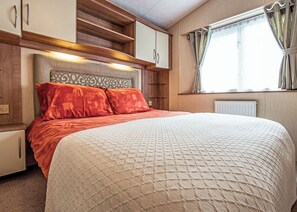 A bedroom with a bed, a headboard, a window with curtains, and built-in wooden cabinets. - 12ft-wide caravan with one double with en suite W. (Tenbury Wells)