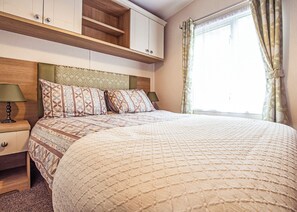 A bedroom with a bed, nightstands, a wooden headboard, a window with curtains, and built-in cabinets. - 12ft-wide caravan with one double with en suite W. (Tenbury Wells)