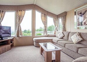 A living room with a sofa, ottoman, glass coffee table, television, and large windows with curtains. - 12ft-wide caravan with one double with en suite W. (Tenbury Wells)