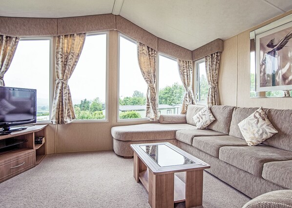 A living room with a sofa, ottoman, glass coffee table, television, and large windows with curtains.