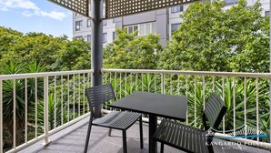 A balcony with two black tables and chairs, surrounded by a white railing, with a view of lush greenery and a building in the background. - One Bedroom in the Heart of  Kangaroo Point with Pool & Parking (Kangaroo Point)