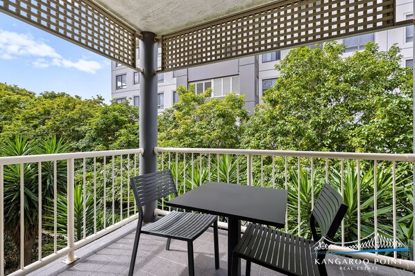 A balcony with two black tables and chairs, surrounded by a white railing, with a view of lush greenery and a building in the background.