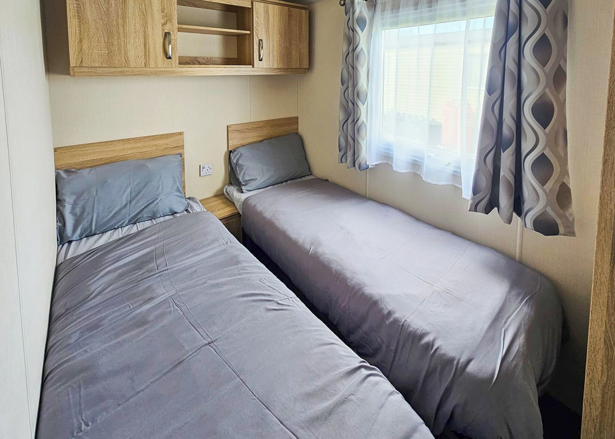 A compact room with two single beds, wooden cabinets above, a window with patterned curtains, and a power outlet on the wall.