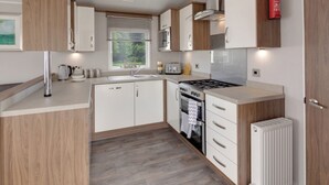 A compact kitchen with a stove, sink, and cabinets, a window with blinds, and a fire extinguisher mounted on the wall.