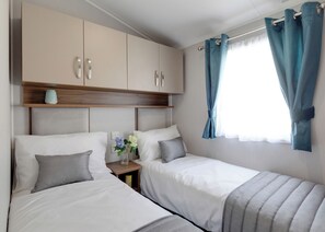 Quarto com duas camas, cabeceira de madeira, mesa de cabeceira com um vaso e janela com cortinas.