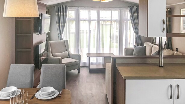 A modern hotel room with a dining area, a sofa, a table set for two, and a large window with curtains.