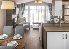 A modern hotel room with a dining area, a sofa, a table set for two, and a large window with curtains. - Spacious and comfortable 12ft-wide caravan with one double with en suite W. (Chapel St Leonards, Skegness)