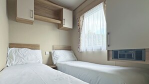 A bedroom with two beds, a window with sheer curtains, a ceiling fan, and a wall-mounted heater.