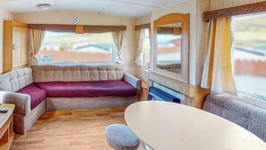 A compact RV interior with a sofa, a dining table, and a small stool. - Comfortable caravan with one double and two twin. (Cross Inn, Nr New Quay)