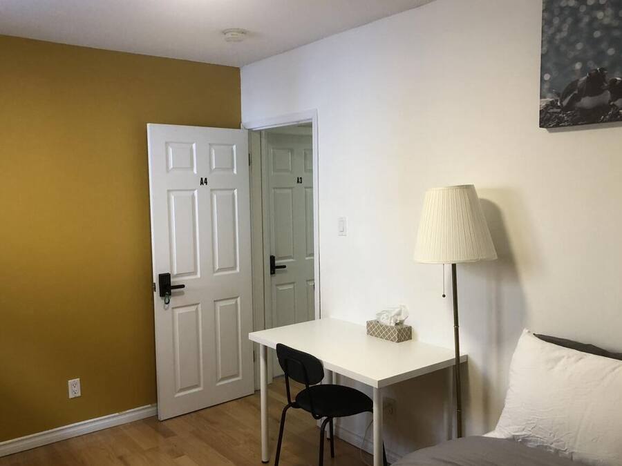 A room with a white desk, a black chair, a bedside table with a lamp, and a bed with a white pillow.