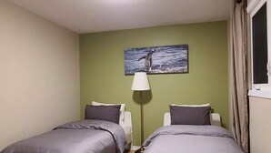 A room with two beds, a floor lamp, a painting of a penguin, and a window with curtains. -  YK Aurora B&B  (Yellowknife)