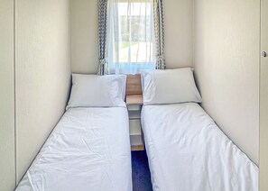 A small, narrow room with two beds, a wooden bench, and a window with patterned curtains. - 2 bedroom accommodation in Newbridge, Nr Yarmouth (Newbridge, Nr Yarmouth)
