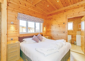 A wooden cabin bedroom with a bed, bedside tables, a window with a curtain, and a bathroom visible through an open door.