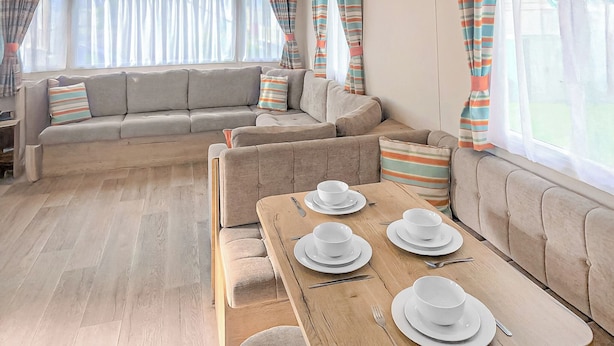 A modern living room with a sofa set, a dining table set for four, and large windows with curtains. - Caravan with one double and two twin. (Chapel St Leonards, Skegness)