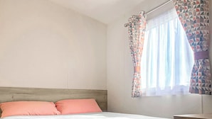 A bedroom with a bed, pillows, a wooden headboard, a window with patterned curtains, and a bedside table. - Caravan with one double and two twin. (Chapel St Leonards, Skegness)