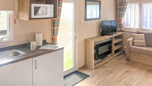 A compact kitchenette with a microwave, a sink, and a small countertop. There is a television on a wooden stand and a sofa with striped cushions. - Caravan with one double and two twin. (Chapel St Leonards, Skegness)