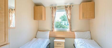 A compact RV bedroom with two beds, a small desk, and a window with curtains.