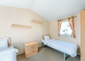 A small, single-room hotel room with a bed, a dresser, a window with curtains, and a wooden shelf. - Fontwell Caravan Plus (Bognor Regis)