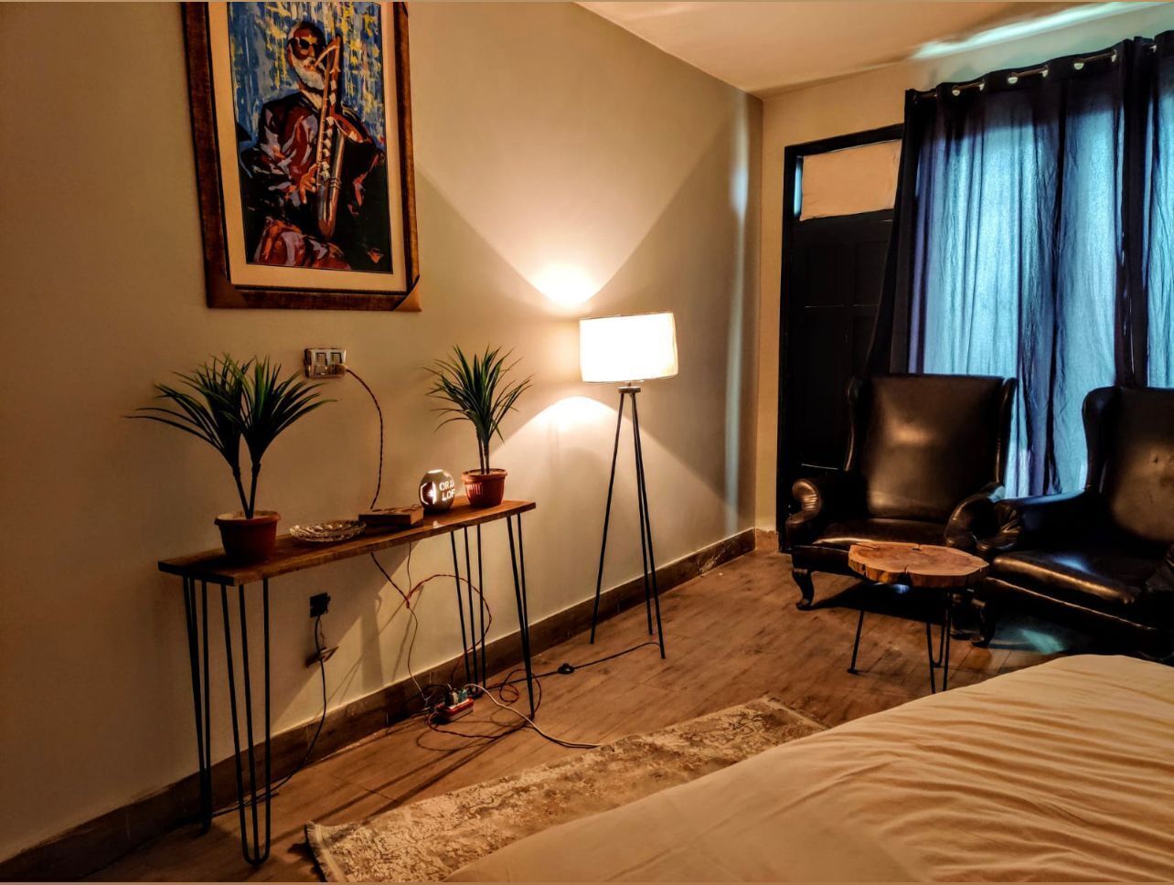 A hotel room with a bed, a desk with two potted plants, a standing lamp, a leather armchair, and a painting on the wall.