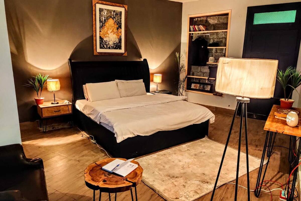 A modern bedroom with a large bed, bedside tables with lamps, a small round table with a book, and a wall-mounted shelf with books and a potted plant.