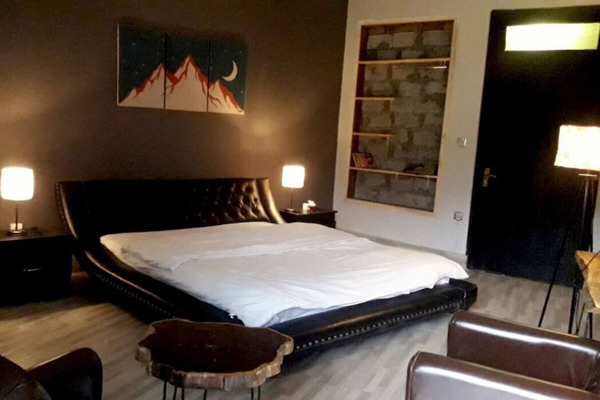 A bedroom with a large bed, two bedside tables, a nightstand, a wall-mounted shelf, and a door with a light fixture.