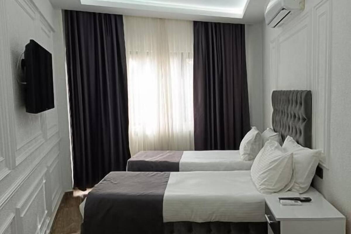 A hotel room with two beds, a flat-screen TV mounted on the wall, a ceiling light, and a window with curtains.