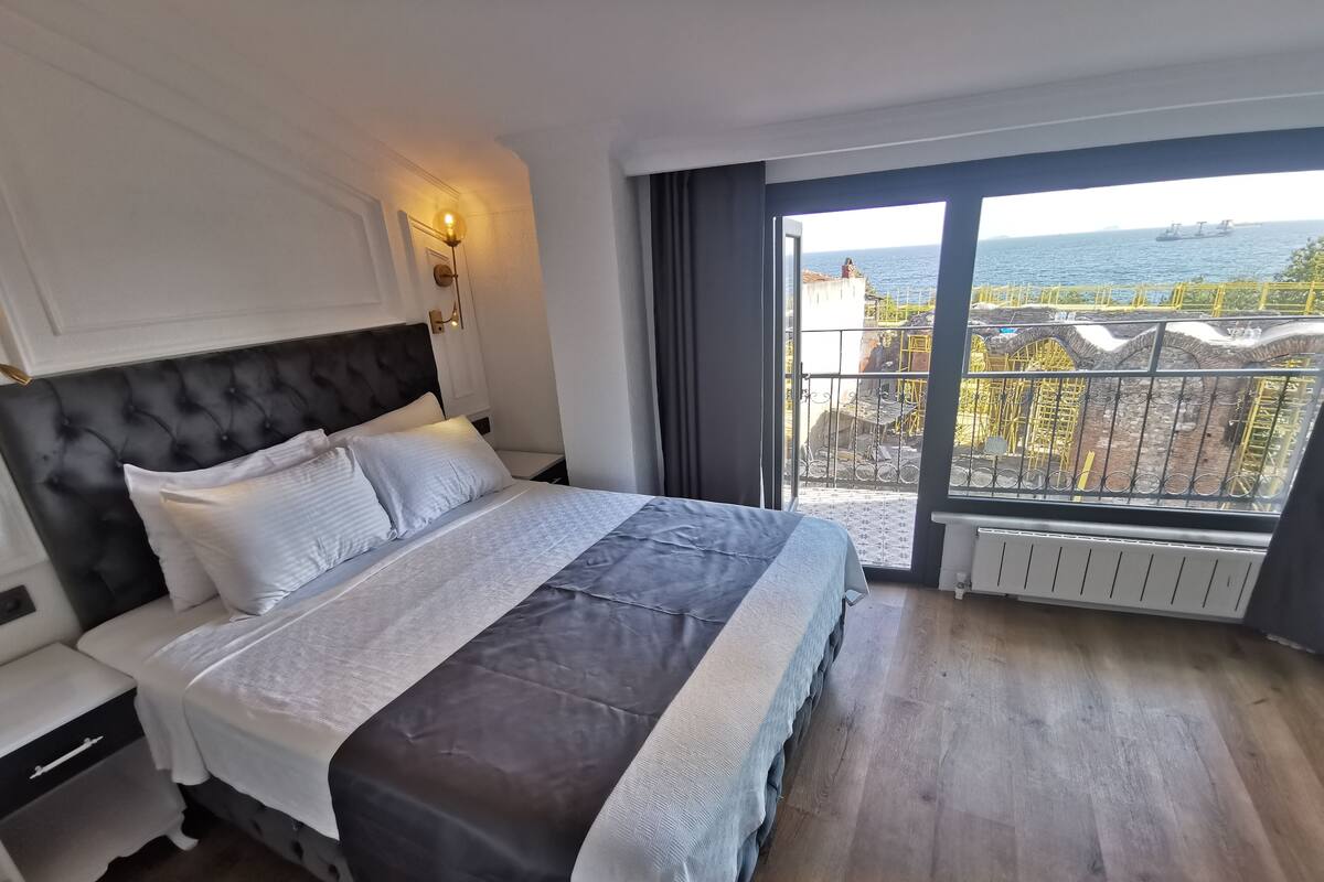 A bedroom with a large bed, a nightstand, a wall-mounted lamp, and a view of the sea through a balcony.