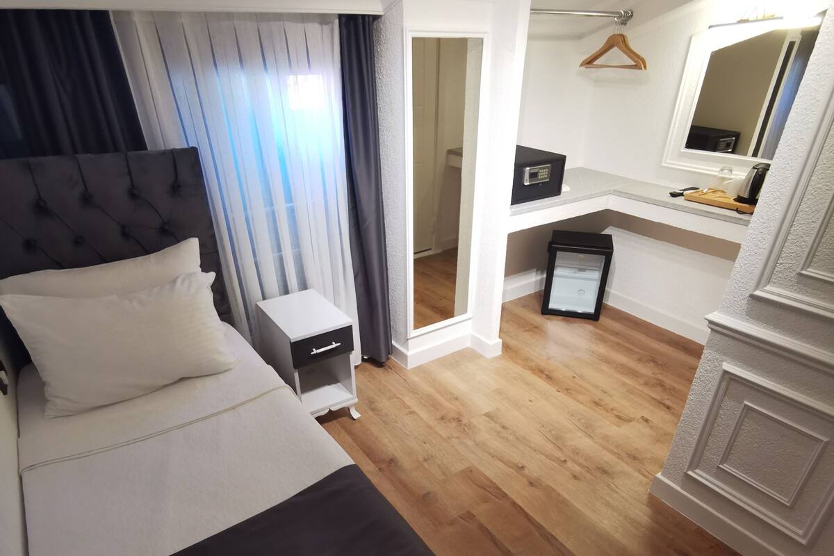 A bedroom with a bed, bedside table, and a small desk with a chair. There is a mirror above the desk and a hanging coat rack.