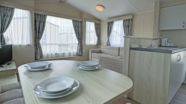 A compact room with a dining table set for four, a sofa, a kitchenette, and large windows with curtains. - Comfortable caravan with one double and one twin. (Mablethorpe, Skegness)