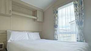 A bedroom with a bed, a nightstand, a wooden headboard, a window with floral curtains, and a view of a house outside. - Comfortable caravan with one double and one twin. (Mablethorpe, Skegness)