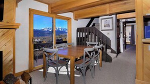 A dining area with a wooden table and chairs, a view of mountains through a large window, and a staircase leading to an upper level. - First Tracks CB, Slopeside, true ski in/out above kids ski school! (Crested Butte)