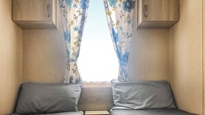 A small, enclosed room with two beds, a window with curtains, and wooden cabinets above the beds.