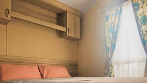 A bedroom with a bed, a wooden headboard, a built-in shelf, and a window with floral curtains.